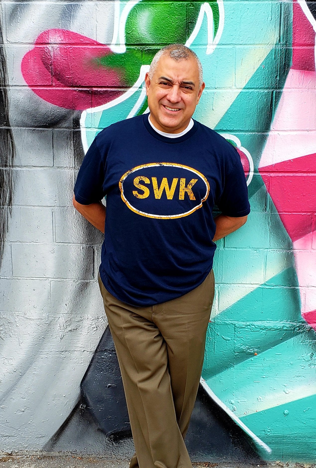 Man standing in-front of a colorful mural wearing Navy colored T-shirt with an orange logo that reads SWK