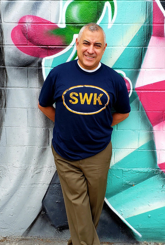 Man standing in-front of a colorful mural wearing Navy colored T-shirt with an orange logo that reads SWK